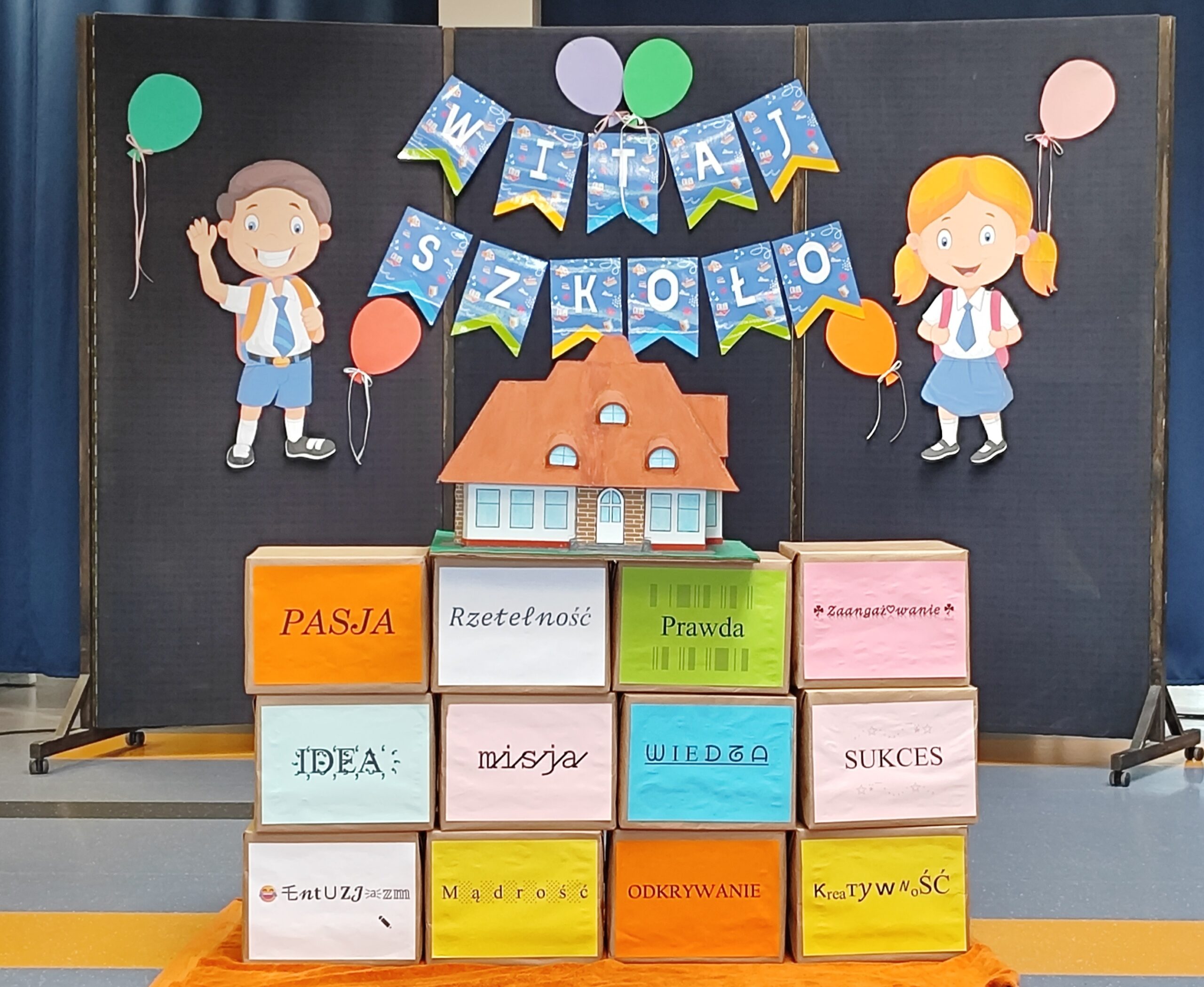 Dekoracja szkolna z napisem „Witaj Szkoło”, postaciami uczniów, balonikami i kolorowymi pudełkami z wartościami: pasja, rzetelność, prawda, zaangażowanie, idea, misja, wiedza, sukces, entuzjazm, mądrość, odkrywanie, kreatywność.