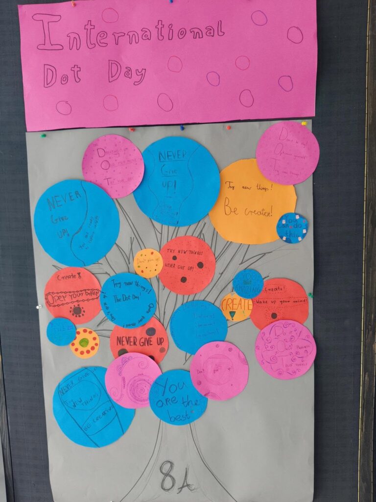 Plakat wykonany przez uczniów klasy 8A z okazji Międzynarodowego Dnia Kropki. Na górze znajduje się różowy napis „International Dot Day” ozdobiony narysowanymi kropkami. Poniżej na szarym tle widnieje rysunek drzewa, którego korona zrobiona jest z kolorowych papierowych kółek w odcieniach niebieskiego, różowego, pomarańczowego i czerwonego. Na każdym kółku zapisano pozytywne i motywujące hasła w języku angielskim, między innymi „Never give up!”, „Be creative!”, „You are amazing!”, „Try new things!” i „You are the best!”.