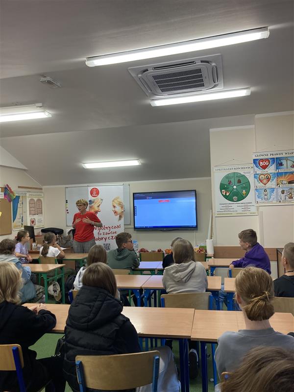 Wolontariusze siedzą w szkolnej klasie podczas spotkania. Przedstawicielka Stwowarzyszenia w czerwonej koszulce stoi przy tablicy multimedialnej i prowadzi zajęcia. Na ścianach wiszą plakaty edukacyjne, a uczniowie siedzą przy ławkach i słuchają.