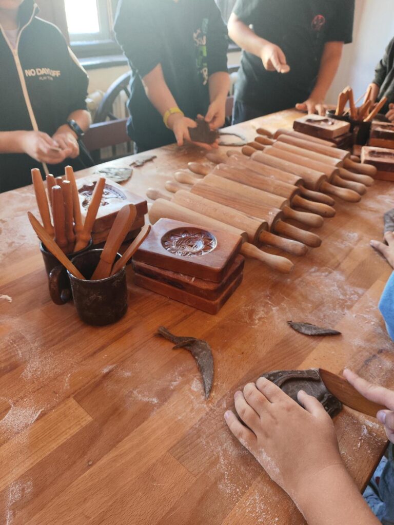 Dzieci uczestniczą w warsztatach pieczenia pierników. Na stole widać wałki do ciasta, drewniane formy z ozdobnymi wzorami oraz narzędzia do modelowania. Dzieci ugniatają i formują ciasto w kształty pierników.