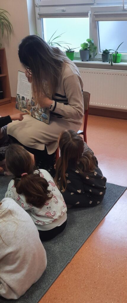 Pani Aleksandra siedzi na krześle i czyta książkę grupie dzieci, które siedzą na dywanie i uważnie słuchają. W tle widać okno z doniczkami z roślinami.