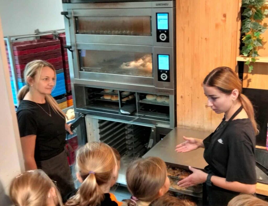 Dzieci obserwują dwie pracownice piekarni przy piecu do wypieku pieczywa. Jedna z kobiet otwiera drzwi pieca, a druga tłumaczy dzieciom proces pieczenia bułek. W tle widać tace z ciastem i gotowe wypieki.