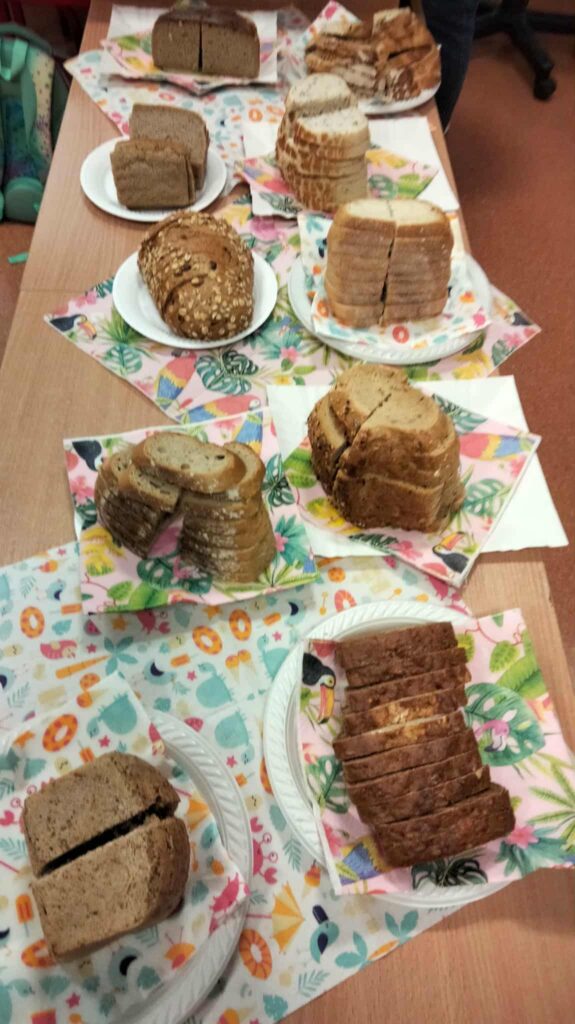 Stół przykryty kolorowymi serwetkami, na którym ustawiono różne rodzaje pokrojonego chleba na talerzach – chleby jasne, ciemne, razowe i z ziarnami, przygotowane do degustacji.