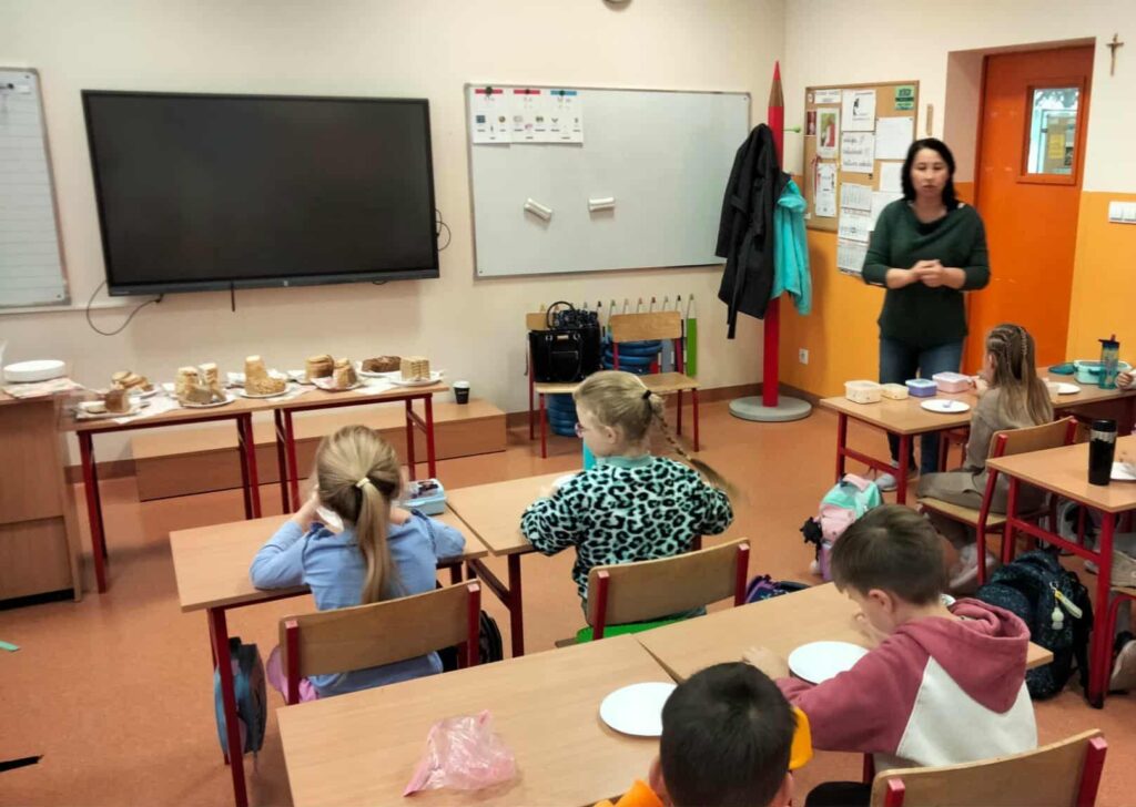 Nauczycielka prowadzi zajęcia w klasie z dziećmi siedzącymi przy ławkach, na stole przed tablicą ułożone są różne rodzaje chleba przygotowane do degustacji.