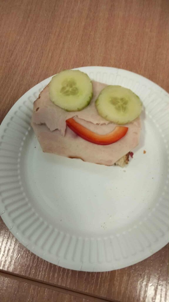 Kanapka ułożona w kształcie uśmiechniętej buzi – oczy z plasterków ogórka, uśmiech z kawałka czerwonej papryki, na kromce chleba z plastrem wędliny.