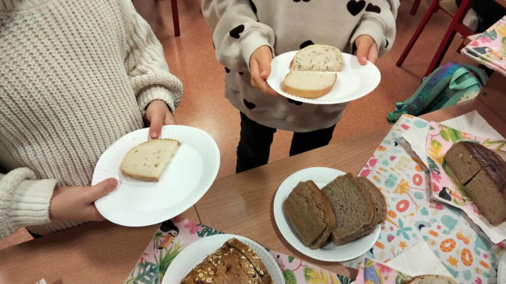 Dwoje dzieci trzyma na papierowych talerzykach kromki różnych rodzajów chleba, stojąc przy stole z ułożonymi do degustacji bochenkami.