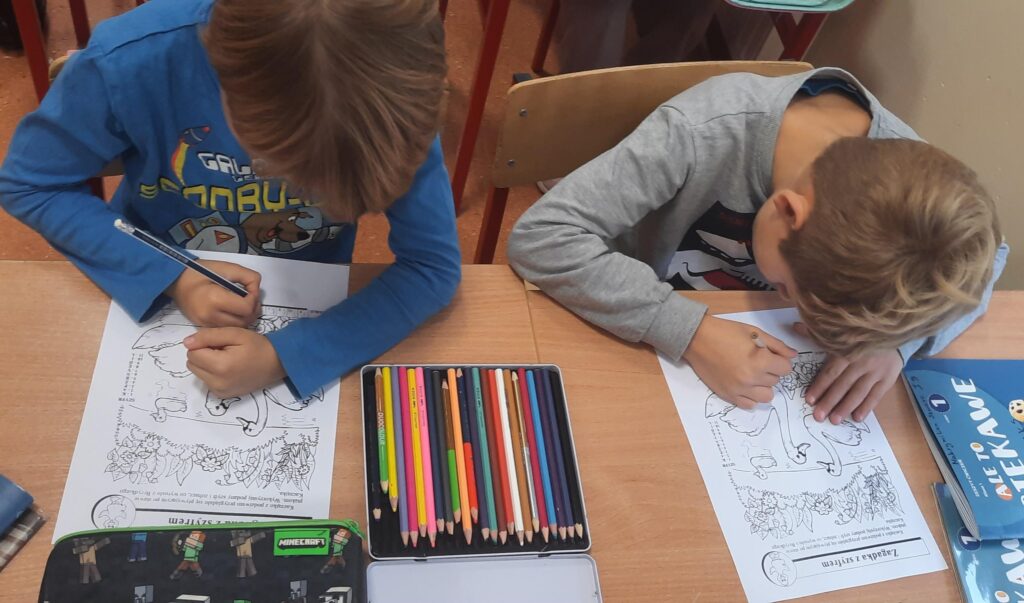 Dwóch chłopców siedzi przy szkolnej ławce i koloruje obrazki przedstawiające sceny z bajki; na stole leży zestaw kolorowych kredek i przybory szkolne.