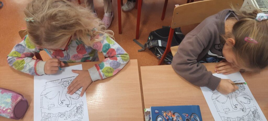 Dwie dziewczynki siedzą przy szkolnej ławce i koloruje obrazki przedstawiające sceny z bajki.