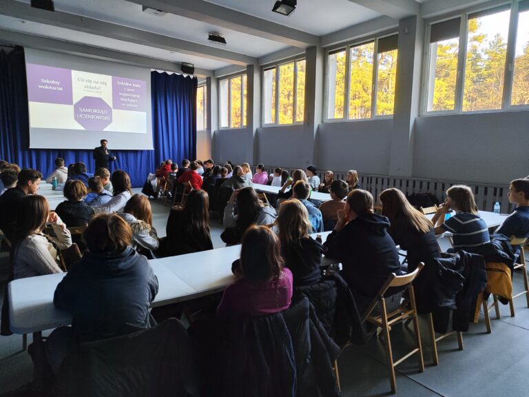 Uczniowie siedzą w auli szkolnej i słuchają wystąpienia działacza