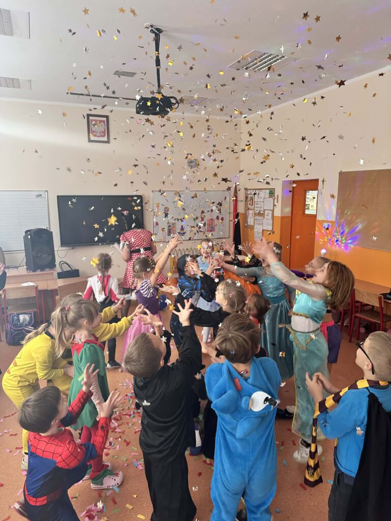 W szkolnej klasie grupa dzieci w kolorowych, bajkowych przebraniach uczestniczy w zabawie. Dzieci stoją w kręgu z wyciągniętymi rękami, próbując złapać złote i srebrne konfetti opadające z sufitu. W tle widać ławki, tablicę, dekoracje i osobę prowadzącą zabawę.
