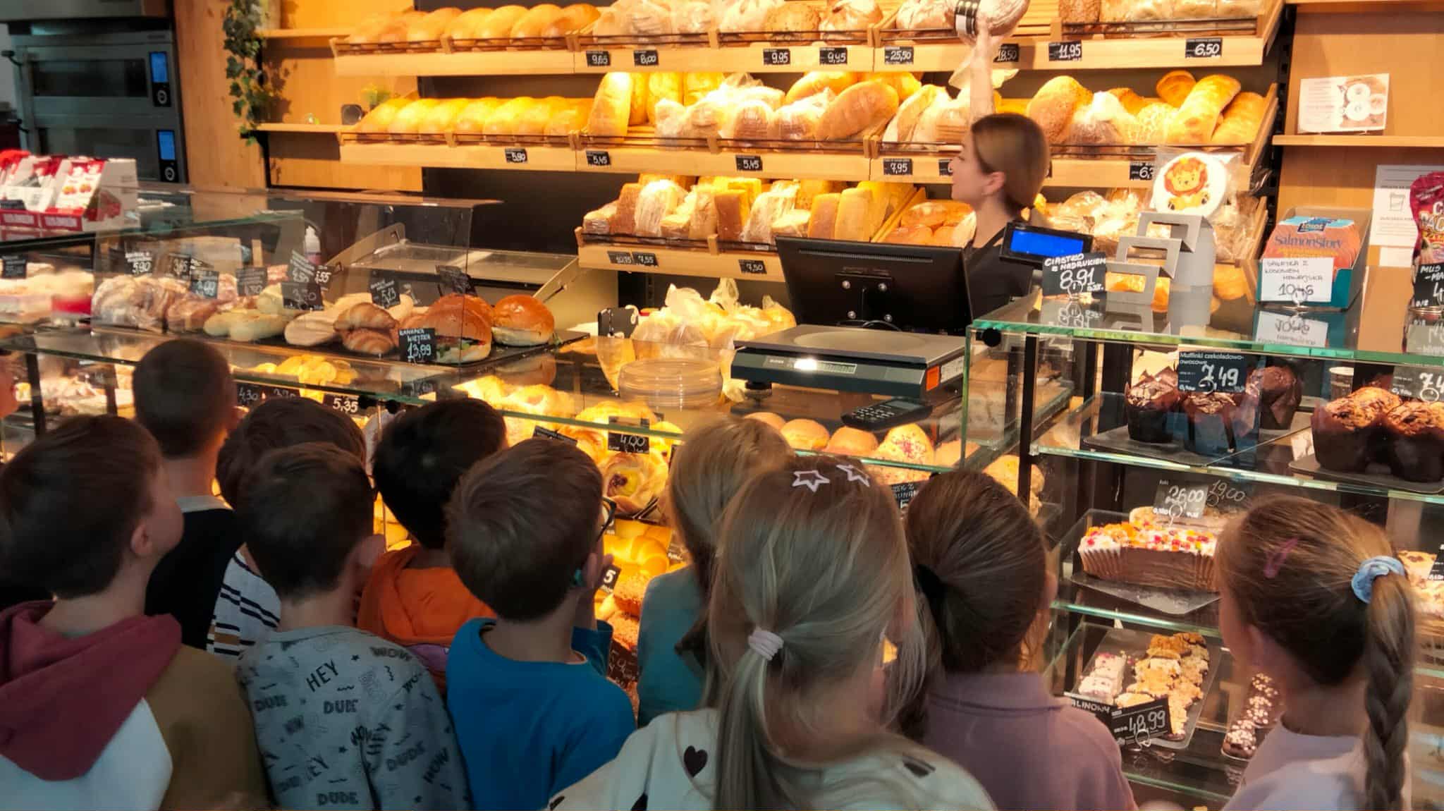 Grupa dzieci stoi przed ladą w piekarni, oglądając różne rodzaje pieczywa i słodkich wypieków. Za ladą stoi sprzedawczyni, która opowiada o produktach. Na półkach widać chleby, bułki i ciasta.