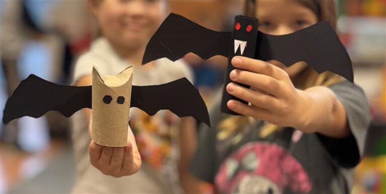 Dwoje dzieci trzyma papierowe nietoperze wykonane z rolek po papierze toaletowym i czarnego papieru, pokazując swoje prace plastyczne
