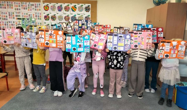 Grupa dzieci stoi w klasie, trzymając przed sobą kolorowe lapbooki, które zasłaniają ich twarze. Każdy lapbook jest ozdobiony wieloma obrazkami i podpisami. W tle widać szkolne plakaty i szafki.