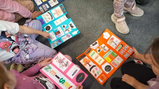 Grupa dzieci siedzi na podłodze i ogląda kolorowe lapbooki z obrazkami i podpisami dotyczącymi różnych tematów; każde dziecko trzyma swoją własną, bogato ozdobioną planszę.