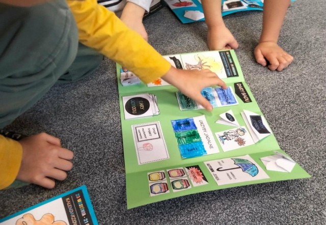 Dzieci siedzą na podłodze i oglądają kolorowy lapbook z obrazkami przedstawiającymi ubrania, pogodę i codzienne przedmioty; jedno z dzieci dotyka elementów na zielonej planszy.