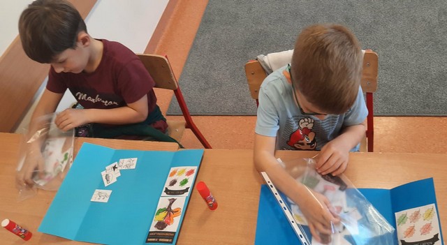 Dwoje dzieci siedzi przy ławce i koloruje edukacyjne karty pracy z ilustracjami, takimi jak ubrania, pogoda i przedmioty codziennego użytku.