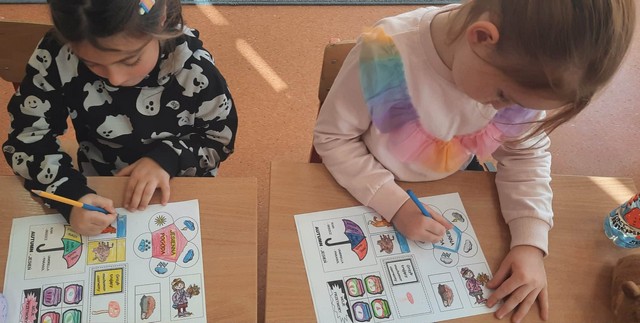 Dwoje dzieci siedzi przy ławce i koloruje edukacyjne karty pracy z ilustracjami, takimi jak ubrania, pogoda i przedmioty codziennego użytku.