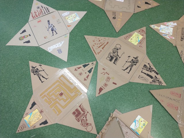 Modele kartonowych piramid z rozkładanym bokiem, ozdobiony egipskimi motywami. Wewnątrz widoczny jest rysunek postaci w stylu starożytnego Egiptu, a na zewnętrznej klapie hieroglify i symbol narysowany na kartonie. Piramida stoi na zielonej podłodze.