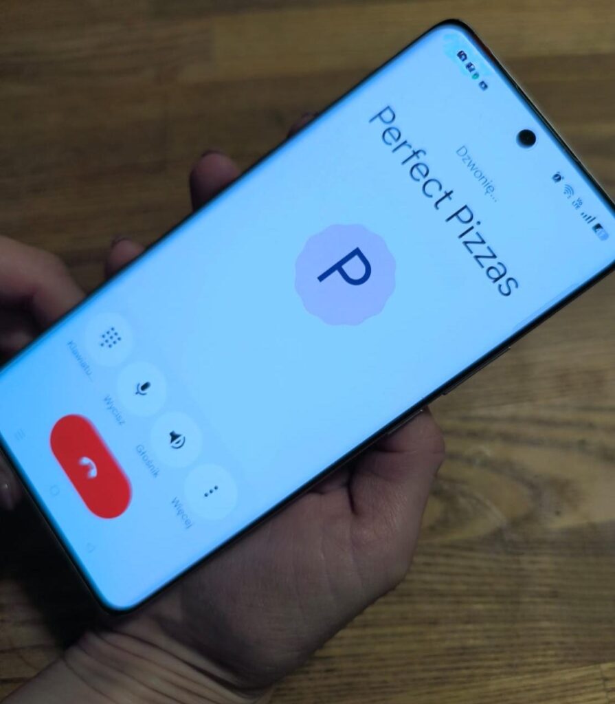 Dłoń trzymająca smartfon, na którego ekranie widać trwające połączenie z kontaktem „Perfect Pizzas”. Na ekranie wyświetlony jest duży inicjał „P” oraz przyciski rozmowy, takie jak wyciszenie, głośnik i zakończenie połączenia.
