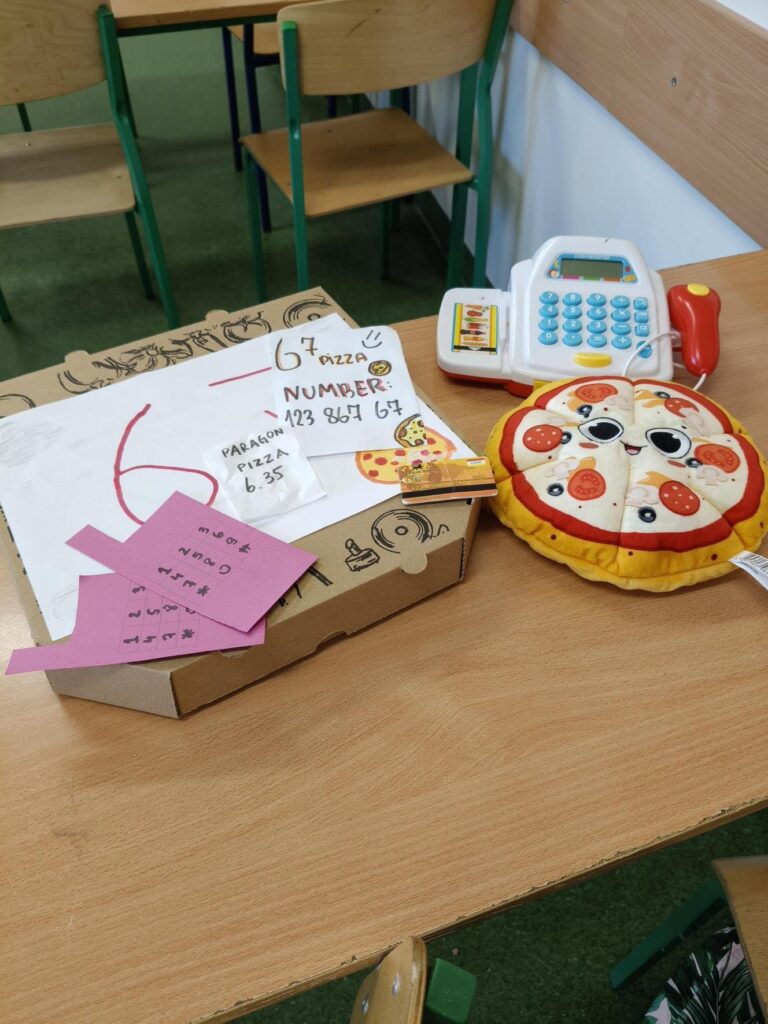 Zestaw zabawkowy „pizzeria” ustawiony na szkolnej ławce: kartonowe pudełko na pizzę z rysunkami, papierowe paragony i karteczki, zabawkowy telefon/kasa z przyciskami, pluszowa pizza w kawałkach oraz plastikowa karta płatnicza.