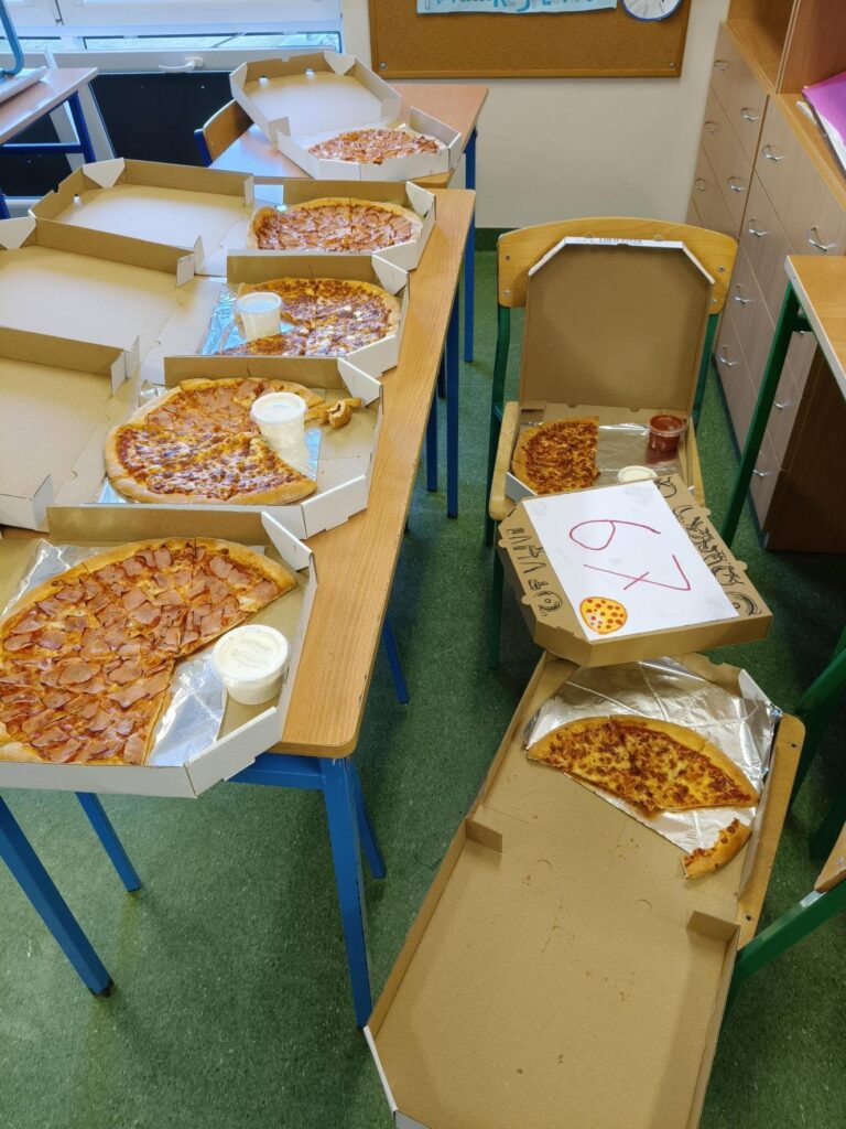 Kilka otwartych pudełek z dużymi pizzami ustawionych na szkolnych ławkach i krzesłach; niektóre pizze są częściowo zjedzone, a obok znajdują się plastikowe pojemniki z sosem. Jedno z pudełek na krześle ma kartkę z dużą czerwoną liczbą „67”.