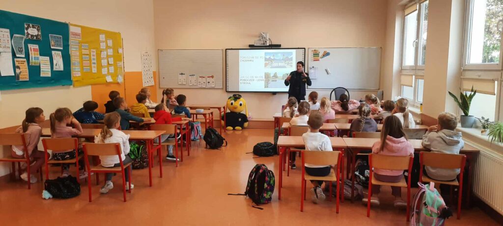 Strażniczka prowadzi zajęcia o bezpieczeństwie drogowym w klasie z uczniami szkoły podstawowej. Dzieci siedzą w ławkach i słuchają prezentacji wyświetlanej na tablicy multimedialnej.
