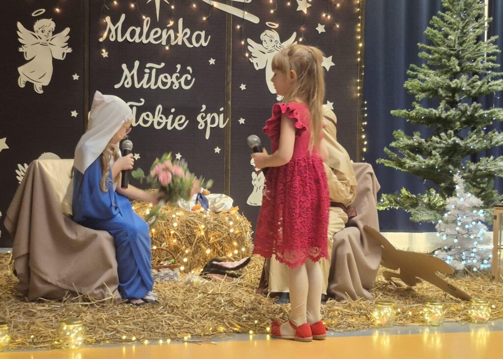 Dzieci występują w szkolnych jasełkach. Dziewczynka w niebieskim stroju Maryi siedzi na scenie na tle czarnej dekoracji z aniołkami, gwiazdami i napisem „Maleńka Miłość Tobie śpi”. Trzyma mikrofon i bukiet różowych kwiatów. Przed nią stoi dziewczynka w czerwonej koronkowej sukience, również z mikrofonem. Obok znajduje się sztuczna choinka ozdobiona światełkami, a cała scena jest udekorowana sianem i lampkami.