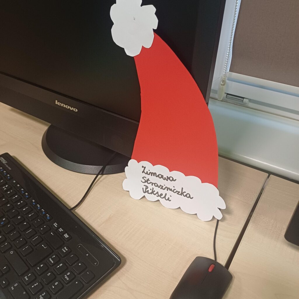 Papierowa czapka Mikołaja z napisem ‘Zimowa Strażniczka Pikseli’ stoi oparta o monitor komputera obok klawiatury i myszy.