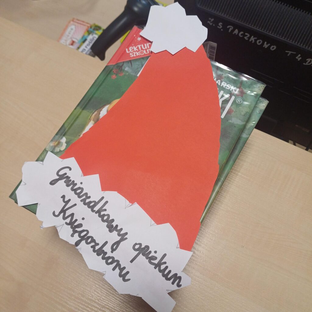 Papierowa czapka Mikołaja z napisem ‘Gwiazd­kowy opiekun Księgozbioru’ leży na stosie szkolnych książek na biurku.”