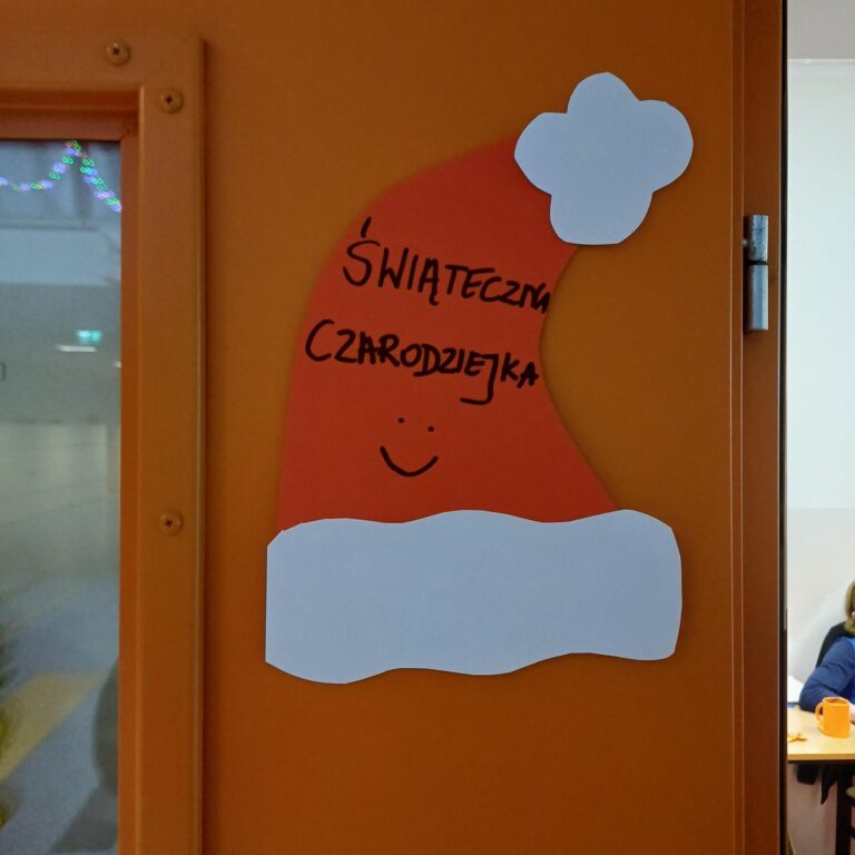 Duża papierowa czapka Mikołaja z napisem ‘Świąteczna Czarodziejka’ wisi na pomarańczowych drzwiach; na czapce narysowano uśmiech.