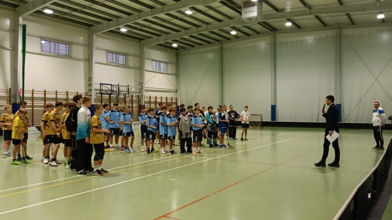 Grupa młodych zawodników w strojach sportowych stoi w szeregu na hali podczas uroczystego rozpoczęcia lub zakończenia turnieju. Po prawej stronie mężczyzna z mikrofonem przemawia do uczestników, a obok niego stoi trener.