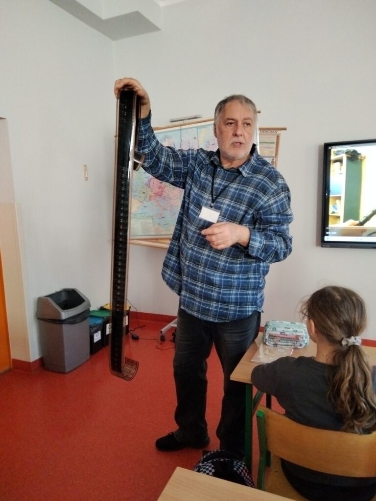 Mężczyzna prowadzący zajęcia w klasie trzyma w ręku długą taśmę filmową i opowiada o niej uczniom siedzącym przy ławkach.
