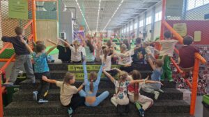 Grupa dzieci siedzących na schodach w strefie trampolin, wspólna zabawa i zajęcia ruchowe w parku rozrywki.