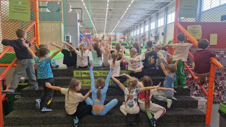 Grupa dzieci siedzących na schodach w strefie trampolin, wspólna zabawa i zajęcia ruchowe w parku rozrywki.