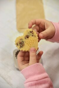 Dziecięce dłonie trzymające naturalny wosk pszczeli w kształcie serca z zatopionymi suszonymi kwiatami.