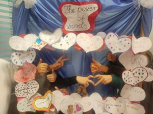 Grupa dzieci stoi za dekoracją z papierowych serc i pokazuje kciuki w górę oraz dłonie ułożone w kształt serca.