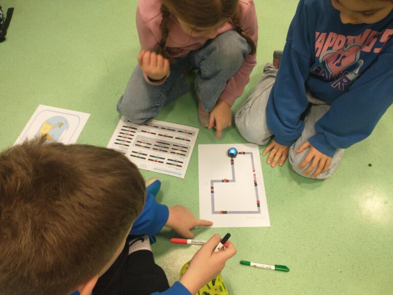 Troje dzieci siedzi na podłodze w klasie i wspólnie pracuje nad zadaniem. Na kartce widoczna jest trasa z kolorowymi polami, po której porusza się mały robot edukacyjny. Uczniowie wskazują elementy na planszy i trzymają w rękach flamastry, analizując rozwiązanie.