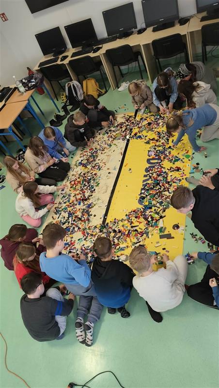 Duża grupa dzieci siedzi w kręgu na podłodze pracowni komputerowej, wspólnie budując i segregując ogromną ilość kolorowych klocków rozsypanych na środku.