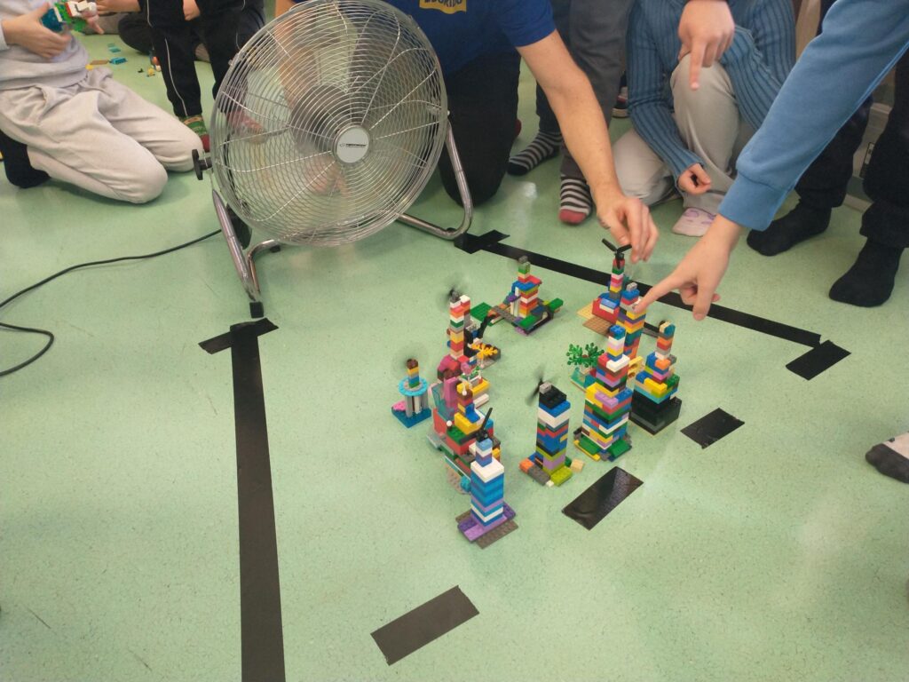 Dzieci siedzą na podłodze wokół makiety zbudowanej z kolorowych klocków. Obok znajduje się duży wentylator skierowany na konstrukcję, a uczniowie wskazują elementy budowli, obserwując efekt doświadczenia.