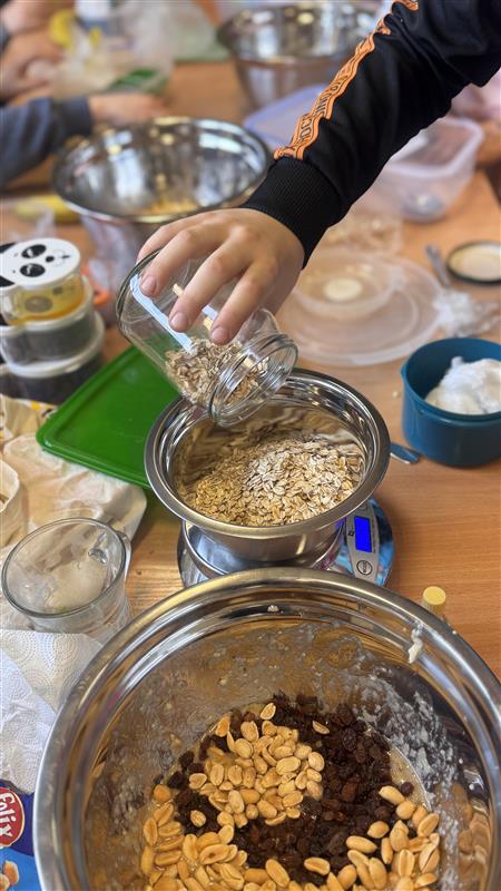 Zbliżenie na rękę wsypującą płatki owsiane z małego słoika do metalowej miski stojącej na wadze kuchennej; wokół na stole znajdują się inne miski i składniki, m.in. orzeszki i rodzynki.