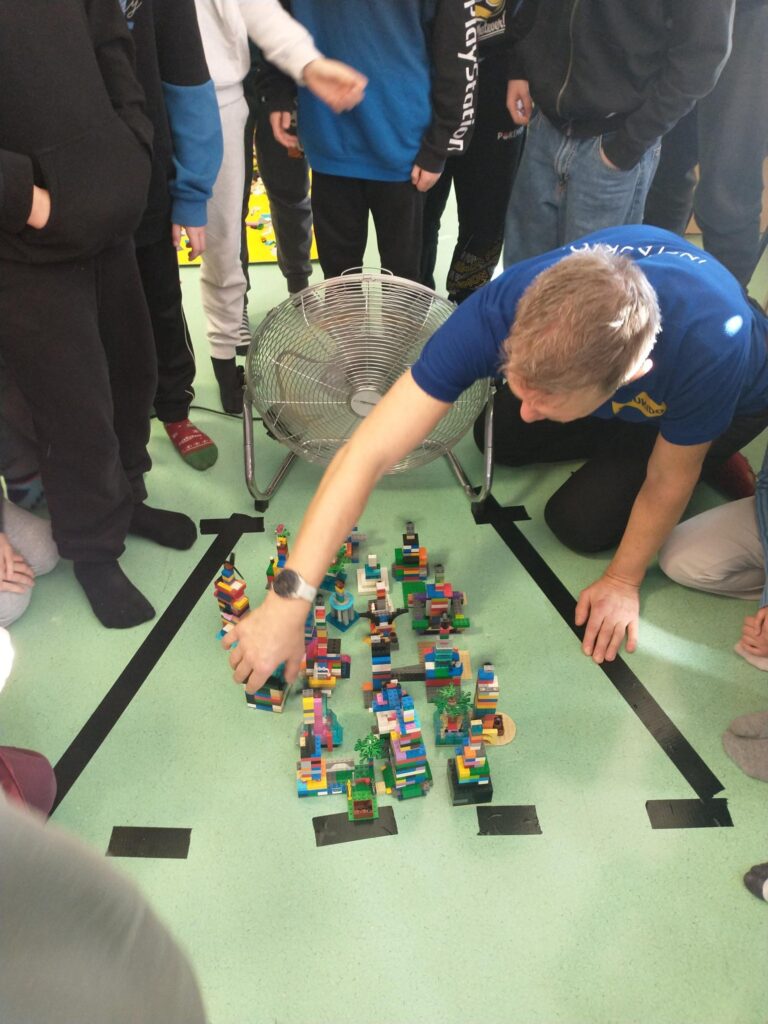 Grupa uczniów otacza makietę miasta z klocków ustawioną na podłodze. Nauczyciel lub opiekun pochyla się i poprawia jeden z elementów, a za nim stoi wentylator używany podczas eksperymentu.