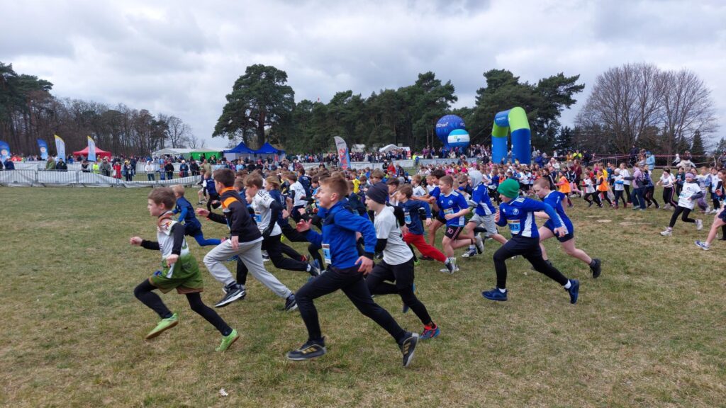 Dzieci uczestniczą w biegu terenowym – duża grupa startuje jednocześnie i biegnie po trawie, a w tle widać tłum kibiców, namioty oraz bramę startową.