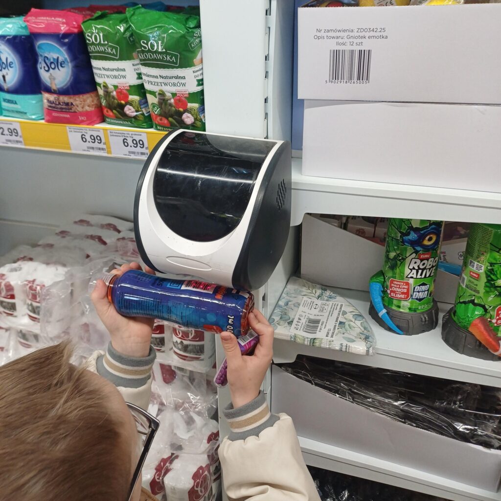 Uczeń w sklepie skanuje kod kreskowy produktu przy czytniku cen. Wokół znajdują się półki z artykułami spożywczymi i zabawkami.