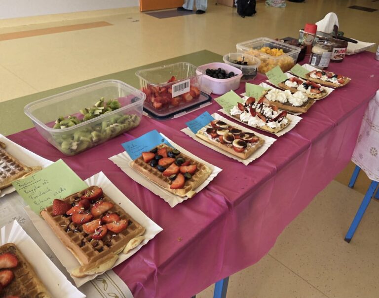 Stół przykryty różowym obrusem, na którym ustawiono kilka gofrów udekorowanych owocami (truskawkami, borówkami, kiwi) i bitą śmietaną. Obok znajdują się plastikowe pojemniki z pokrojonymi owocami oraz dodatki, m.in. Nutella. Przy gofrach leżą karteczki z opisami.