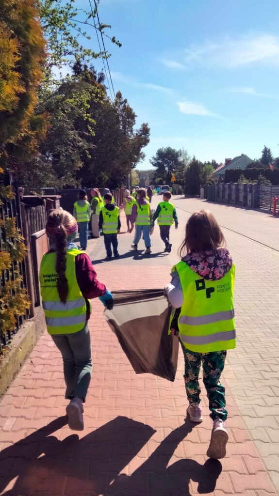 Dzieci w kamizelkach odblaskowych idą chodnikiem z dużym workiem na śmieci. Spacerują osiedlową ulicą w słoneczny dzień w ramach akcji sprzątania okolicy.
