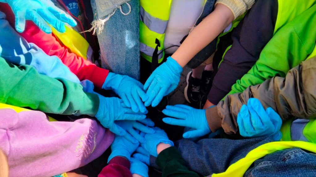 Dzieci stoją w kręgu i wyciągają dłonie w niebieskich rękawiczkach do środka. Gest symbolizuje współpracę i wspólne zaangażowanie podczas akcji ekologicznej.