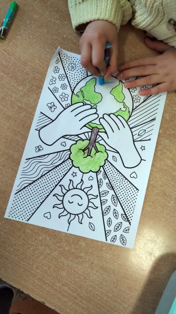 Dziecko koloruje obrazek przedstawiający kulę ziemską trzymaną przez dłonie. Na ilustracji widać także drzewo, słońce, kwiaty i elementy natury. Zdjęcie wykonane na drewnianym stole.