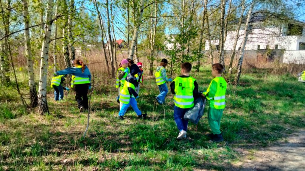 Grupa dzieci w kamizelkach odblaskowych uczestniczy w sprzątaniu terenu zielonego. Dzieci zbierają śmieci do worków wśród drzew i trawy podczas słonecznego dnia.