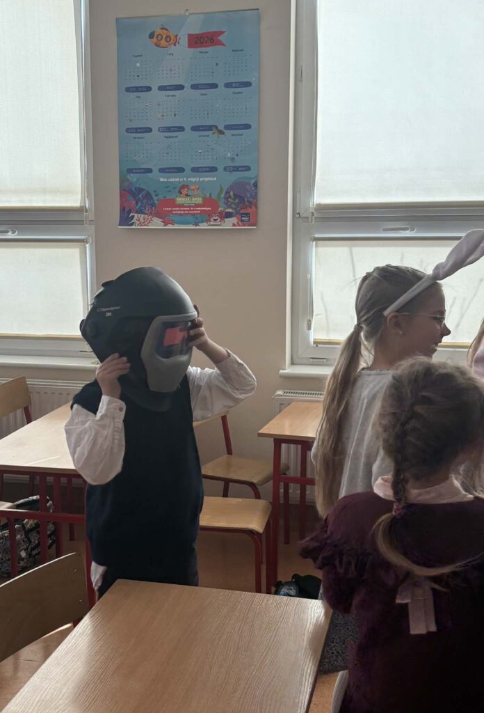 Dziecko stoi w klasie i przymierza dużą przyłbicę spawalniczą. Wokół znajdują się inne dzieci, a w tle widać okna i szkolne ławki.