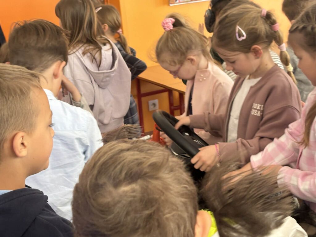 Gromadka dzieci stoi w klasie wokół stolika i z zaciekawieniem ogląda przedmiot przypominający kierownicę. Sala jest kolorowa, a dzieci są skupione i zaangażowane.